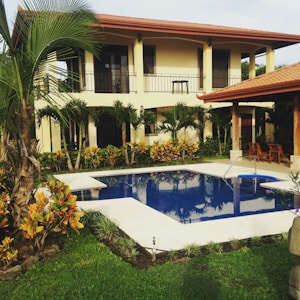 A two-story house with a red-tiled roof and beige walls is surrounded by lush greenery. Palm trees and bushes are planted around the property, which features a clear blue swimming pool with white stone surrounding it. Wooden furniture is placed under a shaded area near the pool.