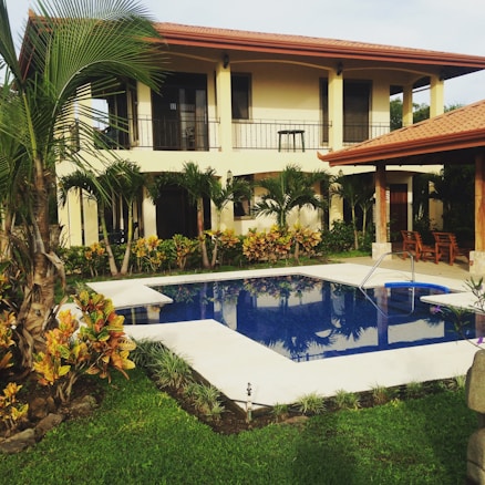 A two-story house with a red-tiled roof and beige walls is surrounded by lush greenery. Palm trees and bushes are planted around the property, which features a clear blue swimming pool with white stone surrounding it. Wooden furniture is placed under a shaded area near the pool.