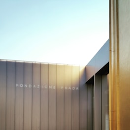 A modern architectural facade with sleek lines and metallic textures, featuring the text 'Fondazione Prada'. The structure is bathed in soft light, with the sky visible in the background.