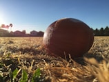 A football is lying on a dry, grassy field under a clear blue sky. The sun is setting or rising in the background, casting a warm glow over the scene. Residential houses and palm trees are visible at the horizon.