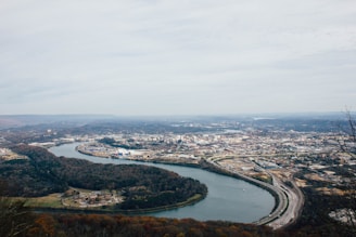 An aerial view of a cityscape with a winding river cutting through the landscape. The city is surrounded by densely wooded areas and rolling hills. Urban structures, roads, and industrial areas are visible, with a highway running parallel to the river.