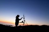 A silhouette of an artist painting on an easel is set against the backdrop of a calming sunset over the ocean. The sky transitions from a warm orange near the horizon to a soothing blue as it moves upward. A small bird is visible flying across the sky.