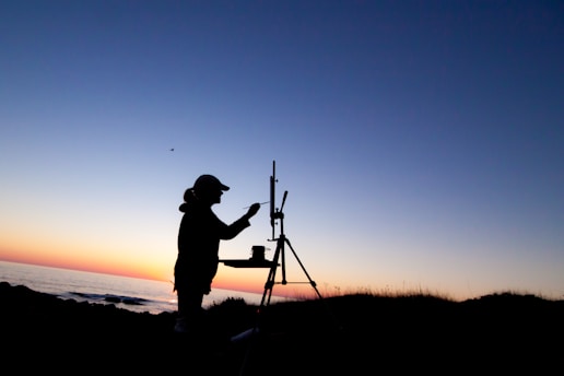 A silhouette of an artist painting on an easel is set against the backdrop of a calming sunset over the ocean. The sky transitions from a warm orange near the horizon to a soothing blue as it moves upward. A small bird is visible flying across the sky.