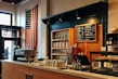 A cozy coffee shop interior featuring an American flag hanging on the wall, a menu board with various coffee options, and a well-organized counter with a coffee machine, bottles, and pastries. The wooden shelves in the background are stocked with jars and bags, while the room has a warm, inviting atmosphere.