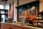 A cozy coffee shop interior featuring an American flag hanging on the wall, a menu board with various coffee options, and a well-organized counter with a coffee machine, bottles, and pastries. The wooden shelves in the background are stocked with jars and bags, while the room has a warm, inviting atmosphere.