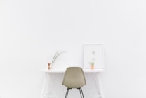 A minimalist workspace with a white desk and a neutral-colored chair placed against a white wall. On the desk, there is a small dried plant in a vase and a picture frame with geometric shapes. The setting conveys simplicity and calmness.