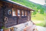 A rustic wooden cabin with a dark wooden exterior wall is decorated with various rustic ornaments. Three small windows with wooden frames allow a view to the outside. A wooden picnic table sits on a stone patio beside the cabin. The surroundings feature a lush green landscape with trees and a grassy hillside in the background.