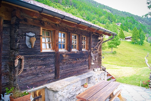 A rustic wooden cabin with a dark wooden exterior wall is decorated with various rustic ornaments. Three small windows with wooden frames allow a view to the outside. A wooden picnic table sits on a stone patio beside the cabin. The surroundings feature a lush green landscape with trees and a grassy hillside in the background.