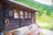 A rustic wooden cabin with a dark wooden exterior wall is decorated with various rustic ornaments. Three small windows with wooden frames allow a view to the outside. A wooden picnic table sits on a stone patio beside the cabin. The surroundings feature a lush green landscape with trees and a grassy hillside in the background.