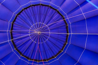 The image is a close-up view of the interior of a hot air balloon as seen from directly below, showing the intricate pattern of the fabric panels. The panels are mostly blue with some white and red ropes creating lines and shapes that add geometric interest to the composition. The central point is a small circular section where the panels converge.