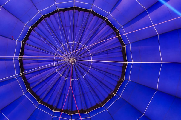 The image is a close-up view of the interior of a hot air balloon as seen from directly below, showing the intricate pattern of the fabric panels. The panels are mostly blue with some white and red ropes creating lines and shapes that add geometric interest to the composition. The central point is a small circular section where the panels converge.
