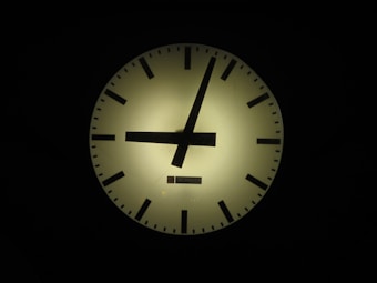 A large, illuminated clock face with black hour and minute markers as well as hands. The clock is set against a dark background, emphasizing its circular shape and glow. The brand name appears at the bottom of the clock face.