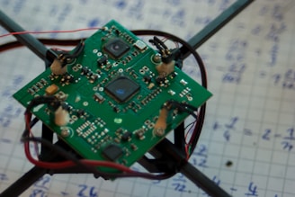 A small green circuit board with various components such as microchips and wiring sits on a graph paper covered in handwritten mathematical equations. The board is mounted on a frame with black arms extending from its corners.