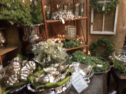 An arrangement of decorative silver items, greenery, and wreaths is displayed in a cozy setting. Various shiny objects like tins and vases are adorned with string lights, while lush green wreaths and plants occupy the shelves and tables. A small sign is visible on a table holding several polished ornaments.