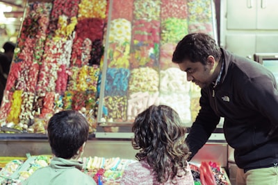 A marketplace scene with a variety of colorful candies displayed in a stall. Two children are eagerly looking at the assortment of sweets while an adult, possibly a vendor or parent, bends over to talk to them.