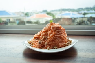 A plate filled with a mound of thinly shaved ice or dessert topped with nuts and a small green garnish, placed on a wooden table in front of a window that reveals a blurry outdoor view.