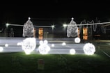 Brightly lit Christmas decorations with spherical light installations and two Christmas trees adorned with lights. The scene is set outdoors at night with a grassy foreground and a building illuminated in the background.