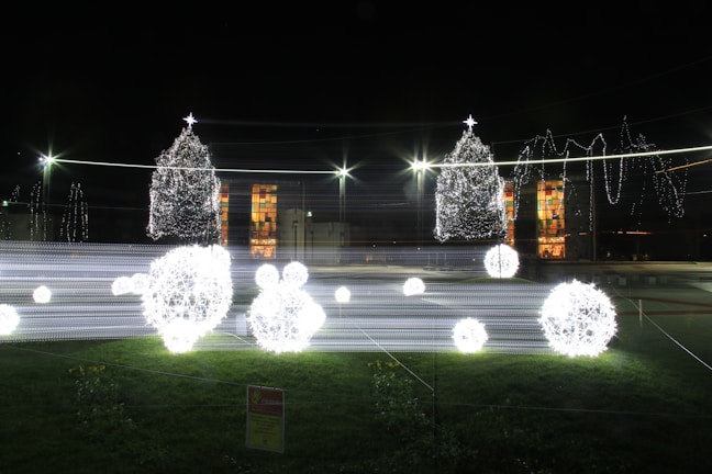Brightly lit Christmas decorations with spherical light installations and two Christmas trees adorned with lights. The scene is set outdoors at night with a grassy foreground and a building illuminated in the background.