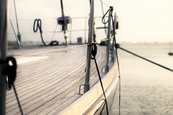 A close-up view of the deck of a yacht with wooden planks running horizontally. The image captures the railing and various ropes secured to it. The background features a calm body of water under a soft, hazy sky.