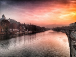 A peaceful river reflecting the soft glow of a vibrant sunset sky. The scene is lined with trees and buildings on either side, and a small dome-topped structure stands out on the left. The atmosphere conveys tranquility with the tranquil water and the horizon fading into gentle hills.