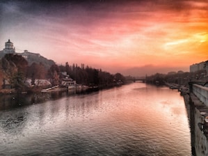 A peaceful river reflecting the soft glow of a vibrant sunset sky. The scene is lined with trees and buildings on either side, and a small dome-topped structure stands out on the left. The atmosphere conveys tranquility with the tranquil water and the horizon fading into gentle hills.
