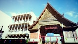 A traditional Thai temple with an intricately designed roof and ornate carvings is positioned next to a modern, multi-story building with balconies. The temple's architecture features detailed gold decorations and a wooden facade. Bright blue sky serves as the backdrop.