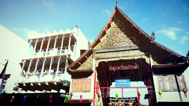 A traditional Thai temple with an intricately designed roof and ornate carvings is positioned next to a modern, multi-story building with balconies. The temple's architecture features detailed gold decorations and a wooden facade. Bright blue sky serves as the backdrop.