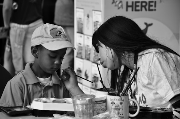 A young woman is painting on the face of a young boy who is wearing a cap. The boy appears focused, while the woman is leaning in to carefully apply paint. Art supplies such as brushes and paint pots are visible on the table in front of them. Other people and informational posters can be seen in the blurred background.