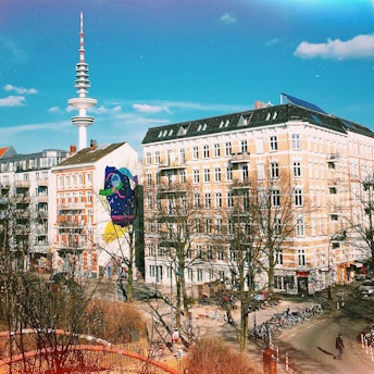 Urban scene featuring a group of traditional European-style buildings with a broadcast tower in the background. The walls of one building display a colorful mural. Leafless trees suggest a late autumn or winter setting, and bicycles are parked along the street. The sky is clear with scattered clouds, casting shadows on the pavement.