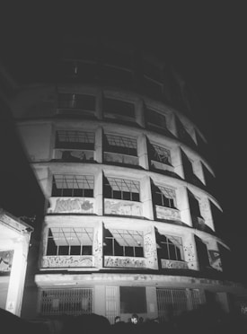 A black and white photograph depicts a multi-story building with graffiti art on its facade. The building appears to be old or possibly abandoned, with intricate iron window grills and visible urban decay. The perspective is upward, emphasizing the structure's height and architectural details.