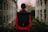 A person wearing a red jacket stands with their back facing the camera, carrying a large black backpack. The setting appears to be an urban environment with two parallel buildings and some greenery, possibly indicating a city street or alley. The sky is overcast, suggesting a cool or autumnal day.