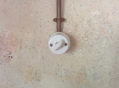 Mounted on a textured wall, a vintage-style ceramic light switch with a circular design is connected to a copper-colored metal conduit. The wall has a rustic, aged appearance with shades of beige and pale brown.