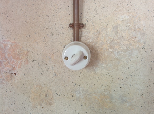 Mounted on a textured wall, a vintage-style ceramic light switch with a circular design is connected to a copper-colored metal conduit. The wall has a rustic, aged appearance with shades of beige and pale brown.