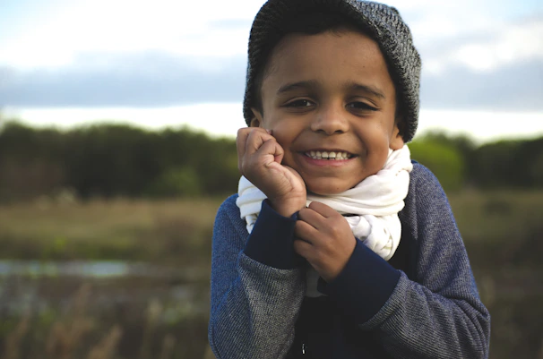 A child wearing a sage green knitted hat, smiling warmly outdoors.