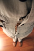 A close-up view of a person wearing a beige linen outfit with a zipper detail and brown shoes. The perspective is looking down at the floor, showcasing the texture of the fabric and the wooden floor beneath.