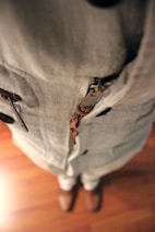 A close-up view of a person wearing a beige linen outfit with a zipper detail and brown shoes. The perspective is looking down at the floor, showcasing the texture of the fabric and the wooden floor beneath.