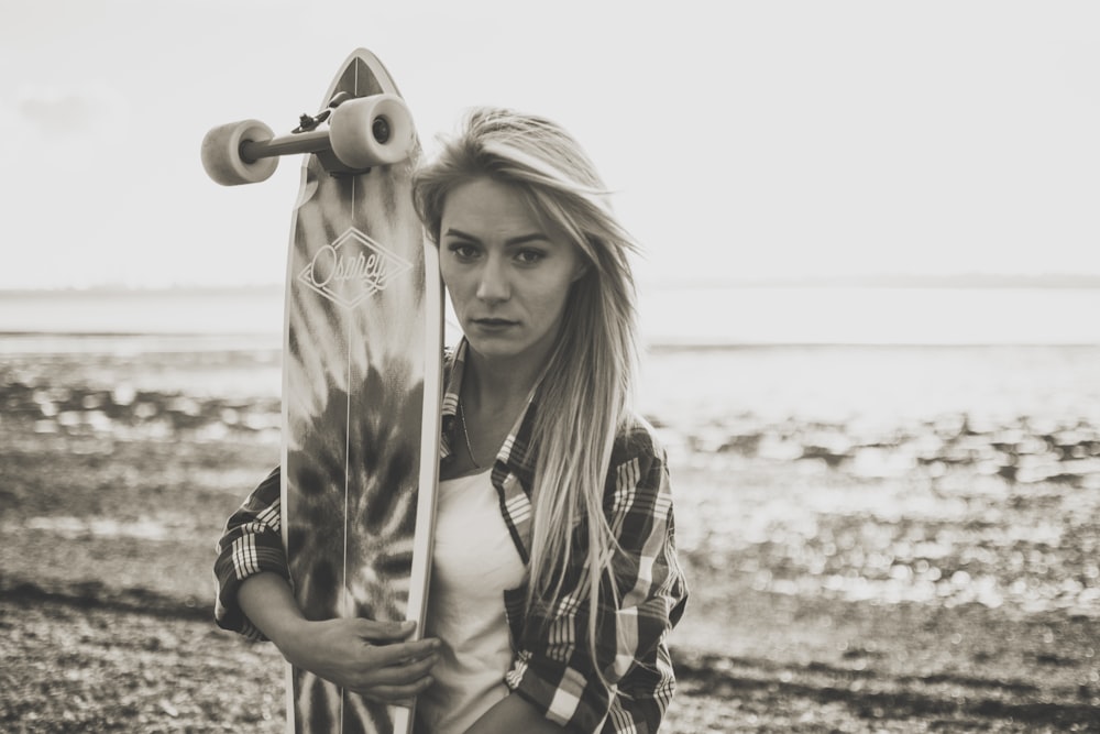 grayscale photography of woman holding a longboard by the beach