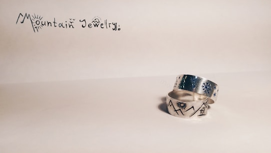 Two silver rings are placed on a light background. One ring has engraved designs of mountains and a sun, while the other features star-like patterns. In the upper left corner, the text reads 'Mountain Jewelry' in a stylized font with decorative elements.