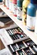 Several paint bottles are neatly arranged on a wooden shelf, with different colors and labels visible. Below the shelf, an open drawer contains various art supplies, including a range of colorful pastels.