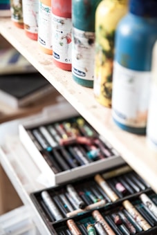 Several paint bottles are neatly arranged on a wooden shelf, with different colors and labels visible. Below the shelf, an open drawer contains various art supplies, including a range of colorful pastels.