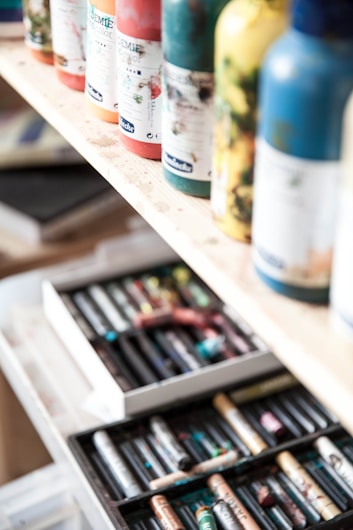 Several paint bottles are neatly arranged on a wooden shelf, with different colors and labels visible. Below the shelf, an open drawer contains various art supplies, including a range of colorful pastels.