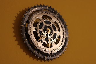 A wall clock crafted from bicycle sprockets, featuring metallic gears prominently displayed against a muted brown background. The hands of the clock are delicate and ornate, contrasting with the rugged appearance of the sprockets.