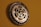 A wall clock crafted from bicycle sprockets, featuring metallic gears prominently displayed against a muted brown background. The hands of the clock are delicate and ornate, contrasting with the rugged appearance of the sprockets.