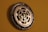 A wall clock crafted from bicycle sprockets, featuring metallic gears prominently displayed against a muted brown background. The hands of the clock are delicate and ornate, contrasting with the rugged appearance of the sprockets.