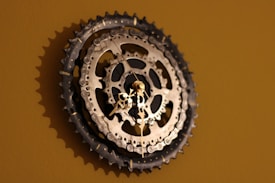 A wall clock crafted from bicycle sprockets, featuring metallic gears prominently displayed against a muted brown background. The hands of the clock are delicate and ornate, contrasting with the rugged appearance of the sprockets.
