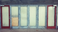 A row of closed doors with frosted glass panels set in a weathered blue frame. The doors are evenly spaced along the front of a building, with two red doors on either end. One of the central panels is covered with a piece of plywood.