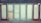 A row of closed doors with frosted glass panels set in a weathered blue frame. The doors are evenly spaced along the front of a building, with two red doors on either end. One of the central panels is covered with a piece of plywood.