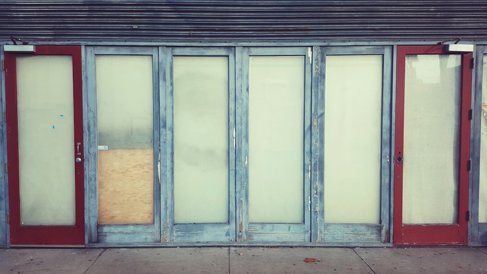 A row of closed doors with frosted glass panels set in a weathered blue frame. The doors are evenly spaced along the front of a building, with two red doors on either end. One of the central panels is covered with a piece of plywood.