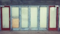 A row of closed doors with frosted glass panels set in a weathered blue frame. The doors are evenly spaced along the front of a building, with two red doors on either end. One of the central panels is covered with a piece of plywood.