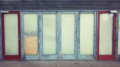 A row of closed doors with frosted glass panels set in a weathered blue frame. The doors are evenly spaced along the front of a building, with two red doors on either end. One of the central panels is covered with a piece of plywood.